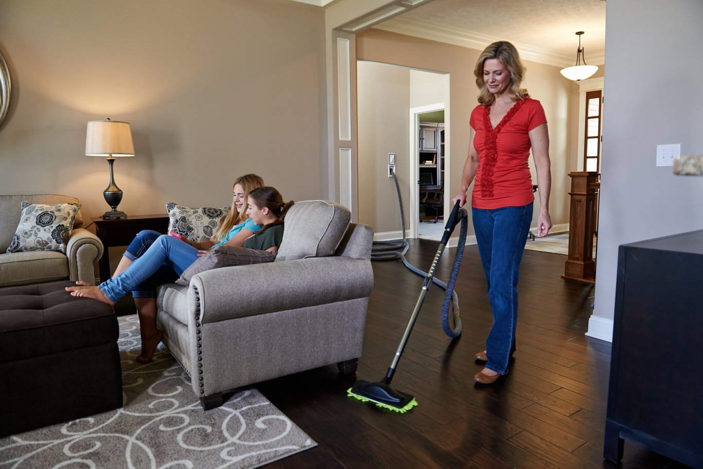 Woman vacuuming a living room with a central vacuum hose while her daughters relax on the sofa