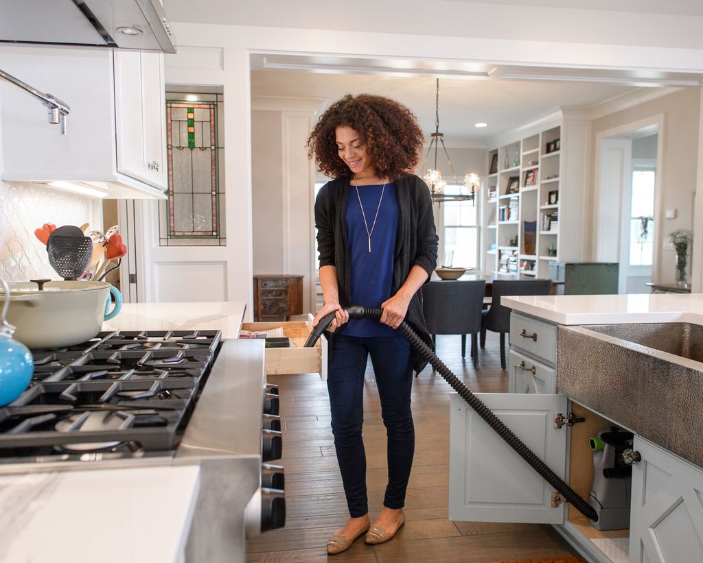 Woman using a retractable under-cabinet central vacuum hose in a bright open-plan kitchen